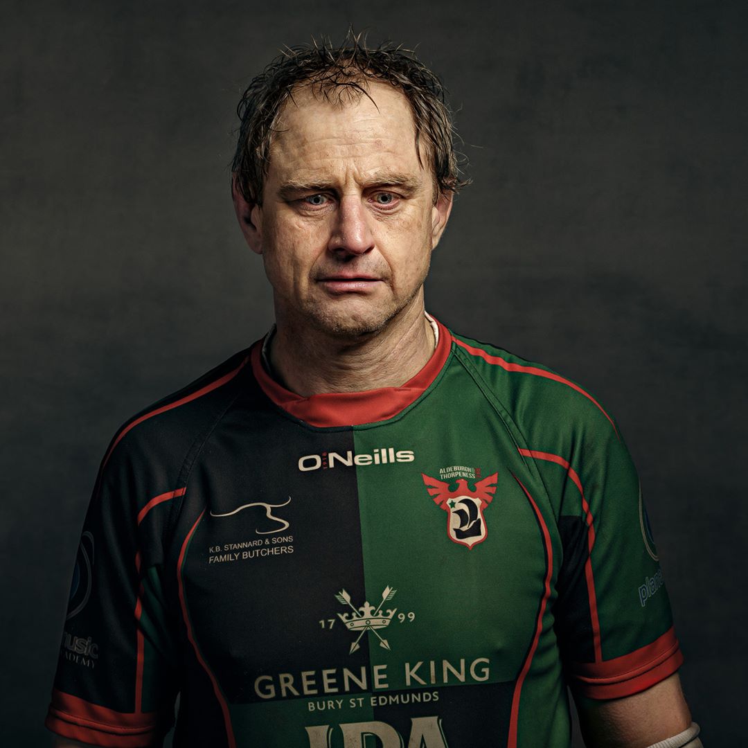A middle-aged man in a green and black rugby shirt with red trim, photographed against a dark grey background.