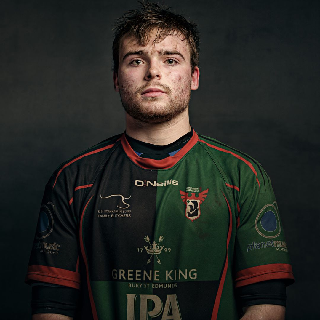 A man in a green and black rugby jersey with red trim looks directly at the camera against a dark grey background.