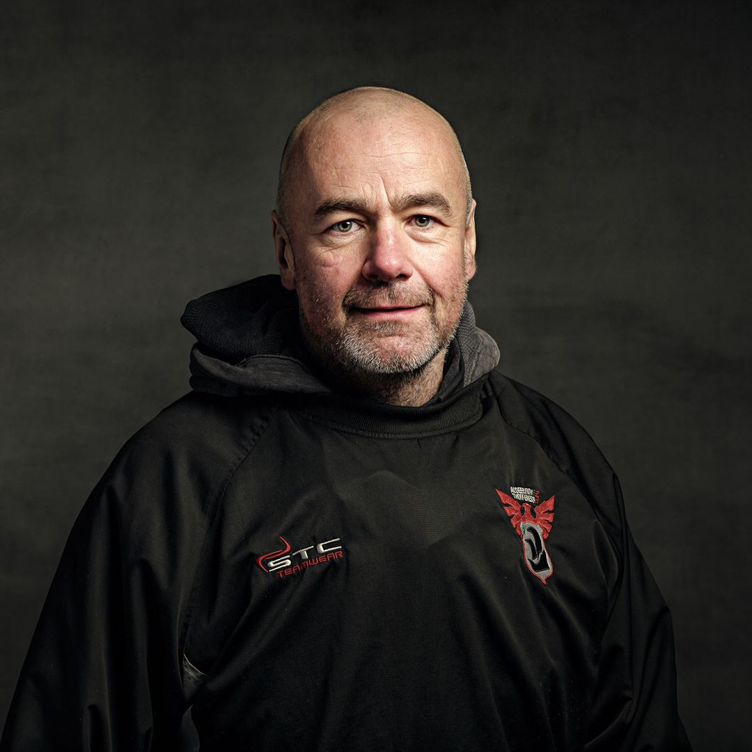 A bald man with a beard wears a black hoodie with red logo details, positioned against a neutral grey background.