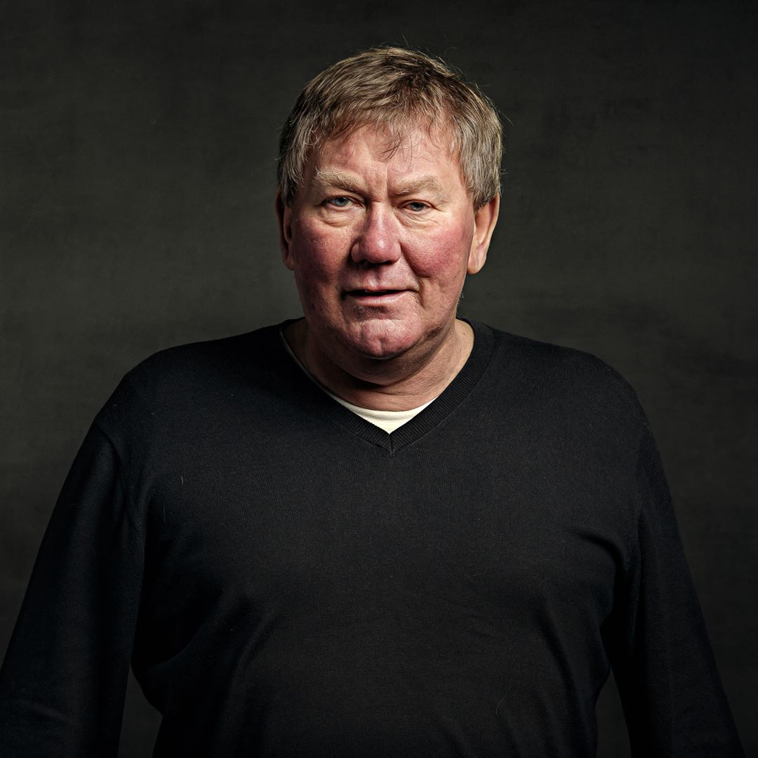 A middle-aged man with grey hair wearing a black v-neck jumper against a dark grey background, looking directly at the camera.
