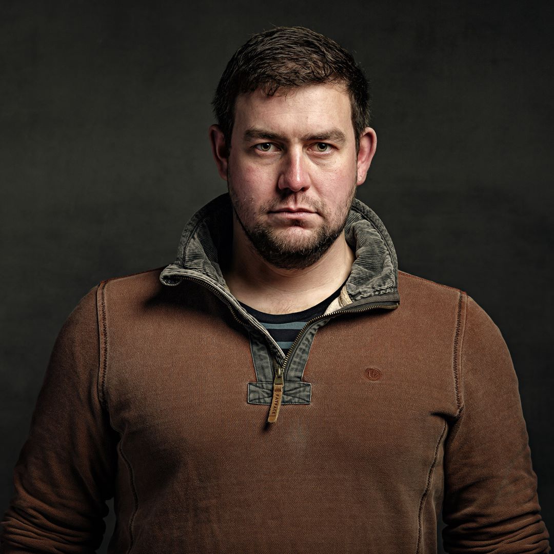 A man with dark hair and beard wearing a brown pullover with grey collar stands against a dark background, looking directly at the camera.