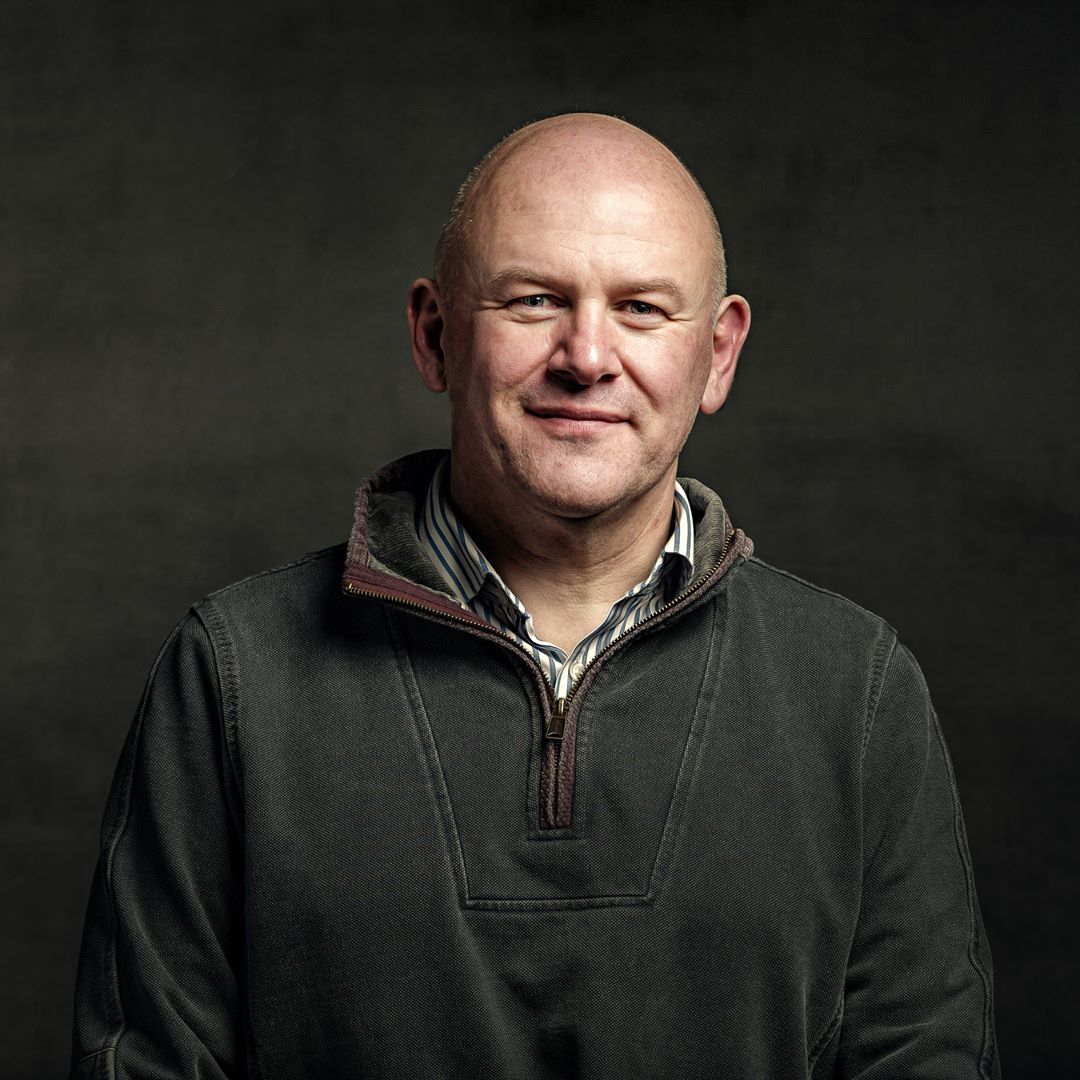 Portrait of a middle-aged man with a bald head, wearing a dark green fleece jacket with a striped shirt underneath, against a dark grey background.