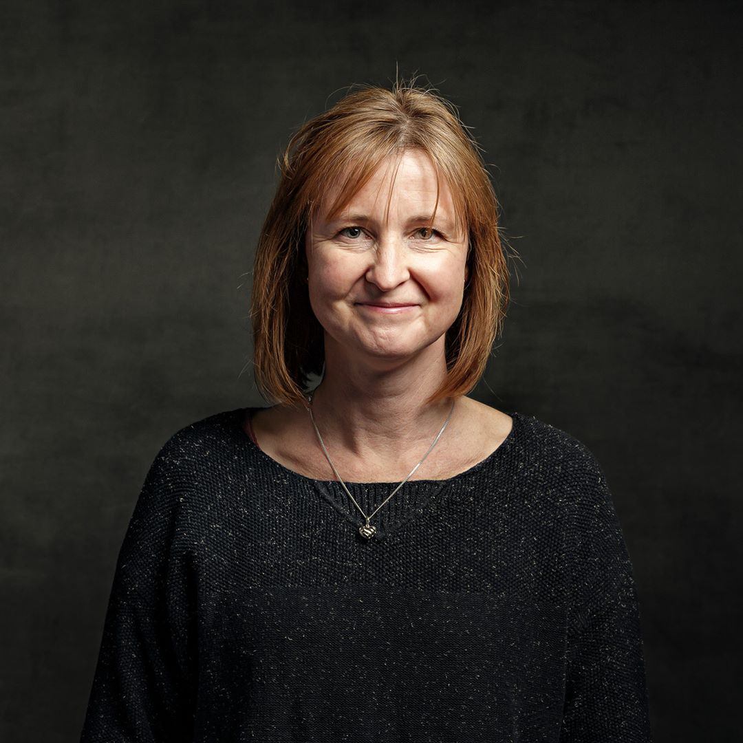 A woman with shoulder-length reddish-brown hair wearing a black textured top and silver necklace, smiling slightly at the camera against a dark grey background.
