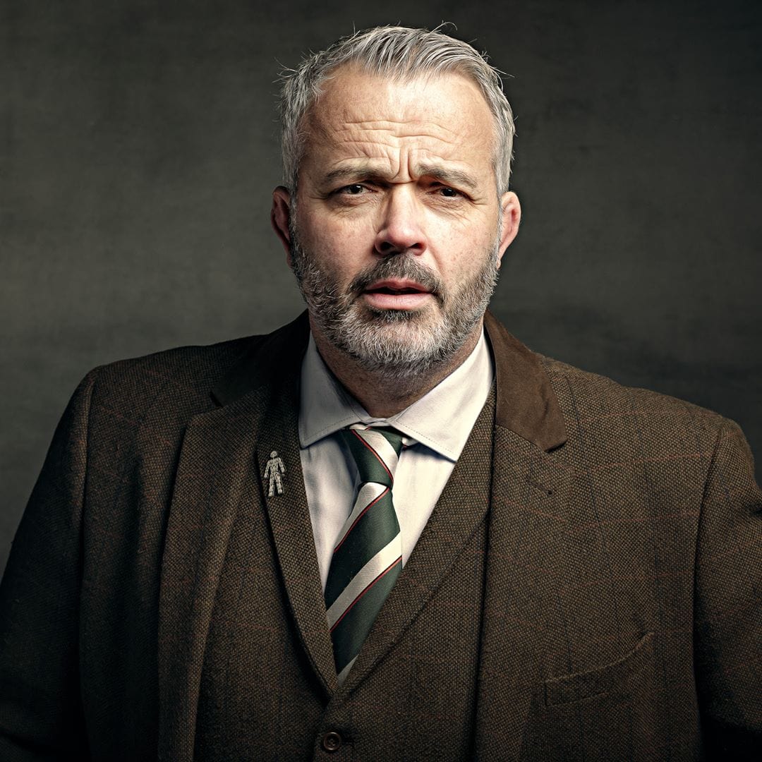 A man with grey hair and beard wears a brown tweed jacket and striped tie against a dark grey background.