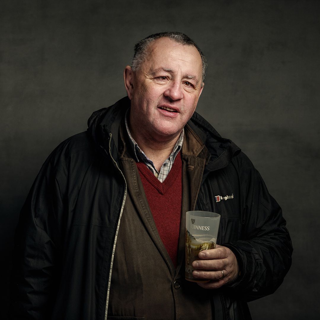 A man in a black jacket holds a Guinness glass against a grey background, looking to the side.