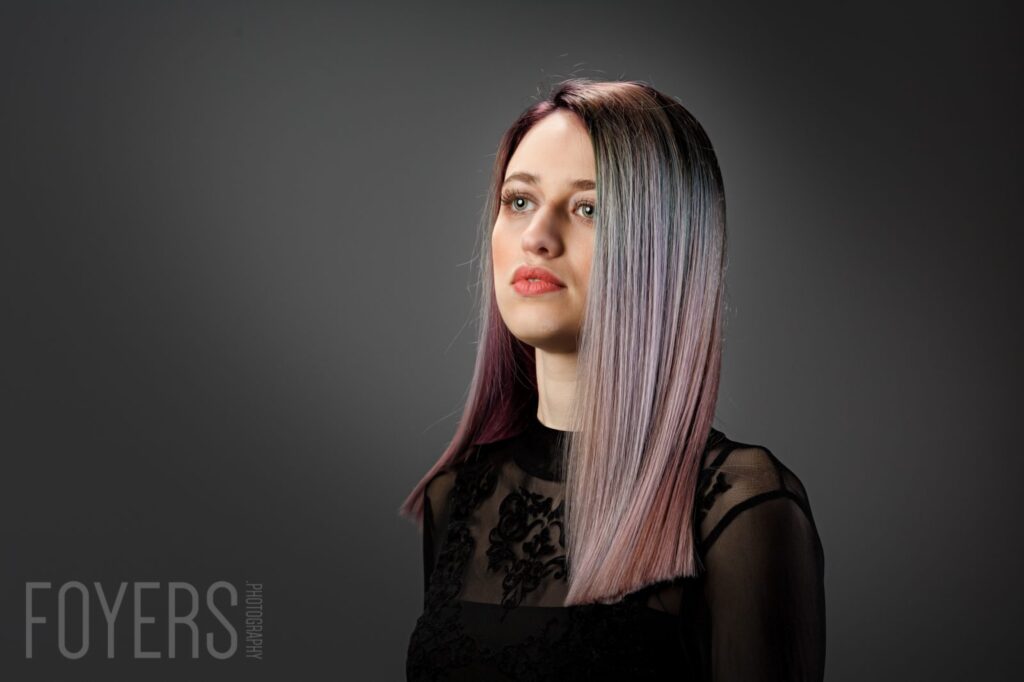A young woman with long straight hair in shades of grey and burgundy faces left against a dark grey background, wearing black.