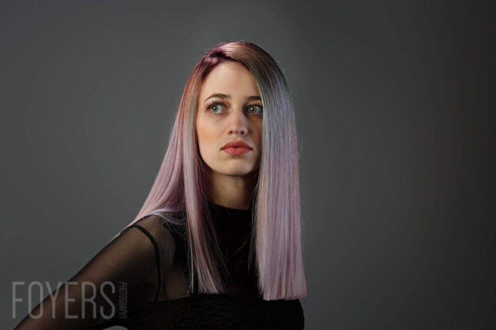 Portrait of a woman with long mauve-pink hair and red lipstick, wearing black, against a grey background.
