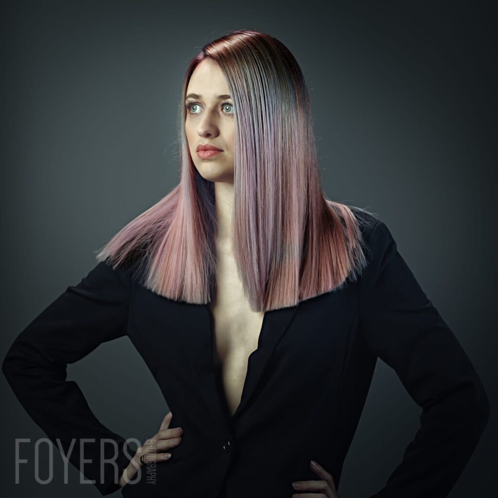 A woman with long straight hair in mauve and pink tones wears a black blazer against a grey background, looking off to the side.