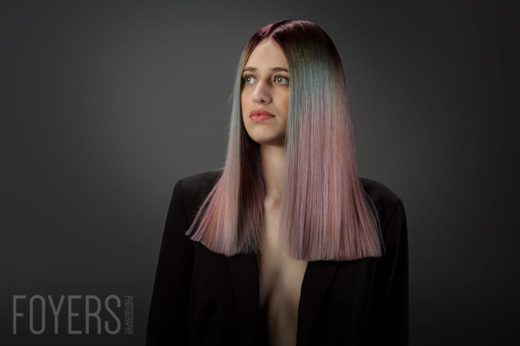 A woman with long straight hair in mauve and black tones, wearing a black blazer, against a grey background. Studio portrait.