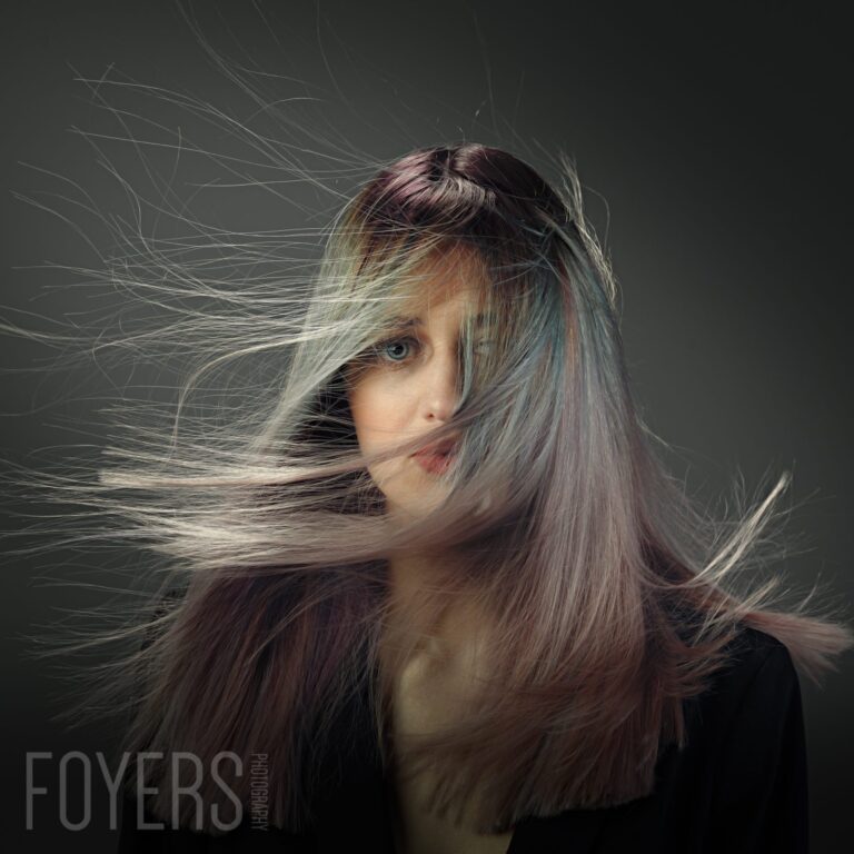 A woman with long dark hair with blonde highlights, her hair blowing across her face, against a grey background.