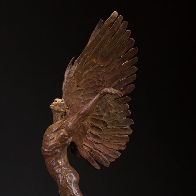 A bronze sculpture of a winged figure against a black background, shown in profile with wings spread upward.