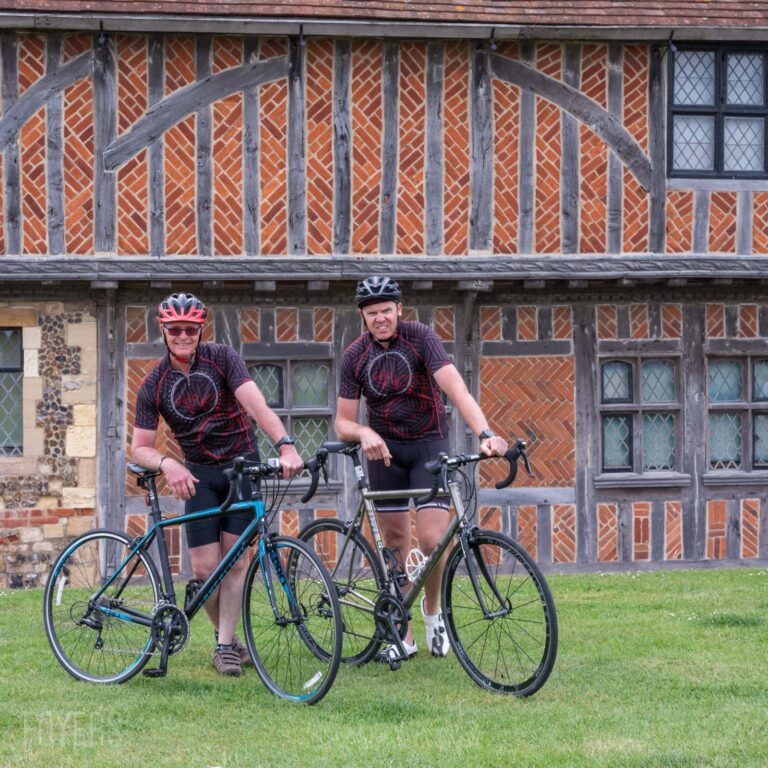 Two cyclists with bikes standing on grass in front of a Tudor-style timber-framed building.