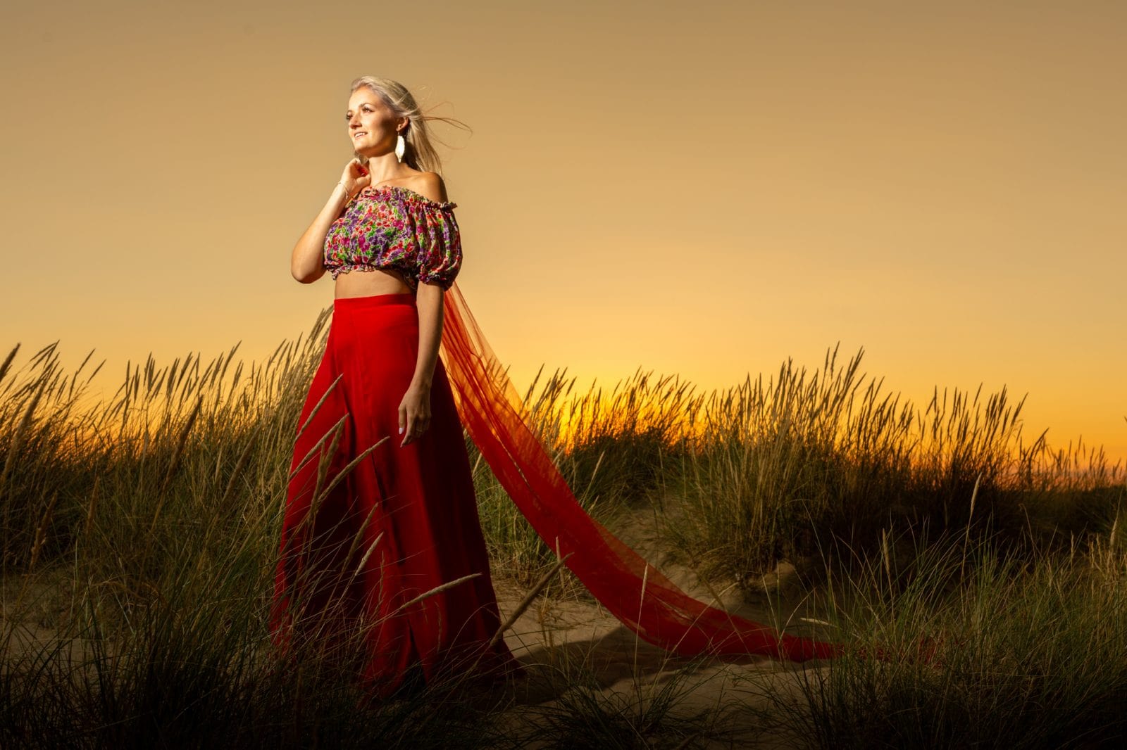 A woman in a red skirt and floral off-shoulder top stands in a sandy field at sunset, looking to the side.