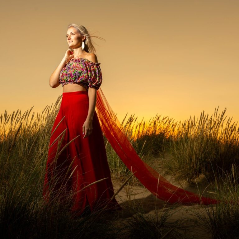 A woman in a colourful off-shoulder top and red skirt stands in a wheat field at sunset, looking to the side.