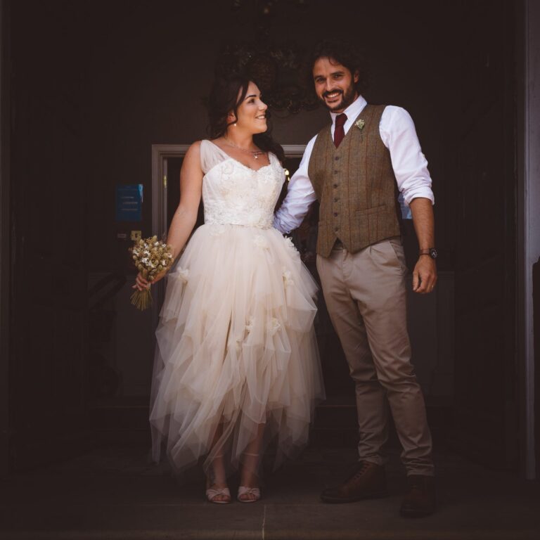 A bride and groom stand together smiling at each other in front of a dark doorway. The bride wears a white tulle dress and holds a small bouquet. The groom wears a brown tweed waistcoat and white shirt.