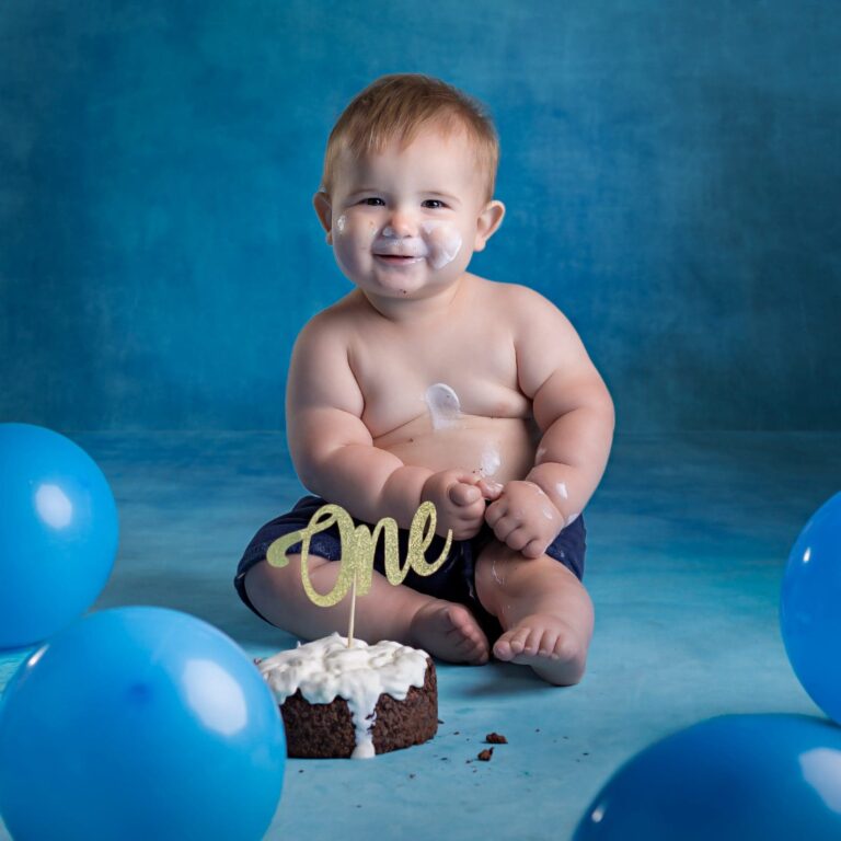 A smiling baby sitting on a blue background with a chocolate cake, one cake topper, and blue balloons surrounding them.