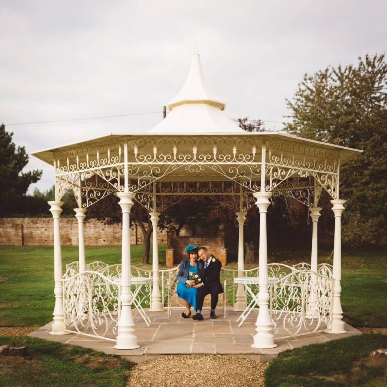 A couple sit together inside an ornate white Victorian gazebo with decorative ironwork, set in a garden with trees and a stone wall.