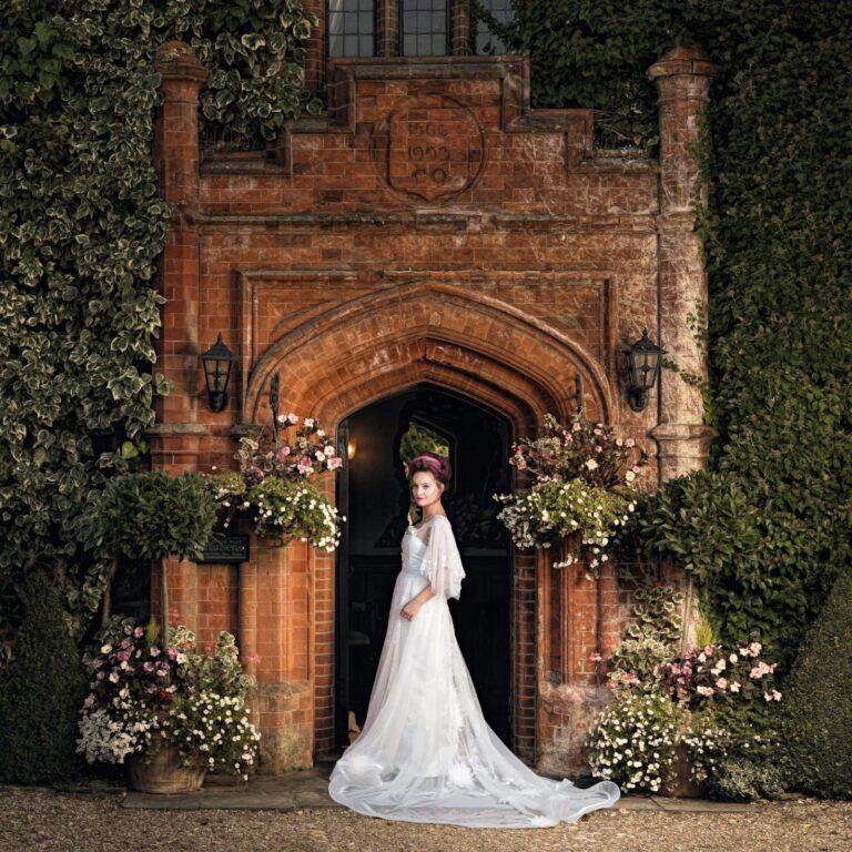 A woman in a white wedding dress and veil stands in front of a brick archway entrance, flanked by flowering planters and ivy.