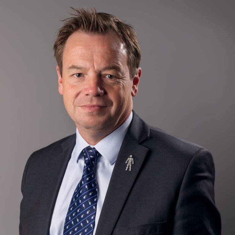 A man in a dark suit and blue patterned tie poses for a professional headshot against a grey background.