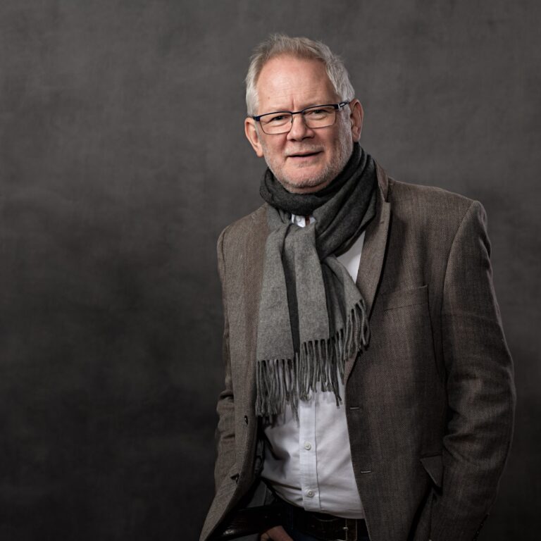 A man with grey hair and glasses wears a brown blazer and grey scarf over a white shirt, standing against a dark grey background.