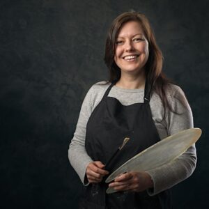 A woman wearing a black apron and grey cardigan holds a wooden spoon and mixing bowl, smiling at the camera against a dark background.