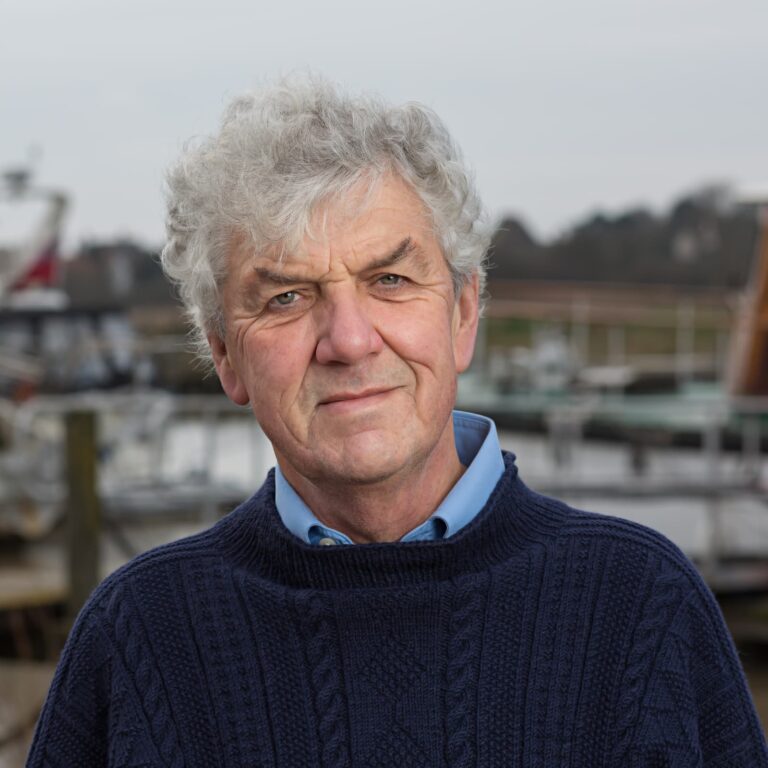 An older man with grey hair wearing a dark cable-knit jumper and light blue shirt, photographed outdoors by a harbour with boats.