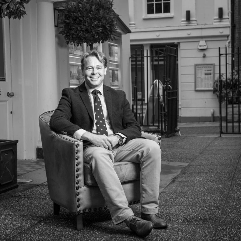 A man in a dark blazer and polka dot tie sits smiling in an upholstered armchair on a paved courtyard outside white buildings.