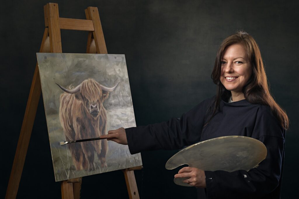 An artist stands beside an easel holding a paintbrush and palette, smiling at the camera. On the easel is her painting of a Highland cattle with long horns.