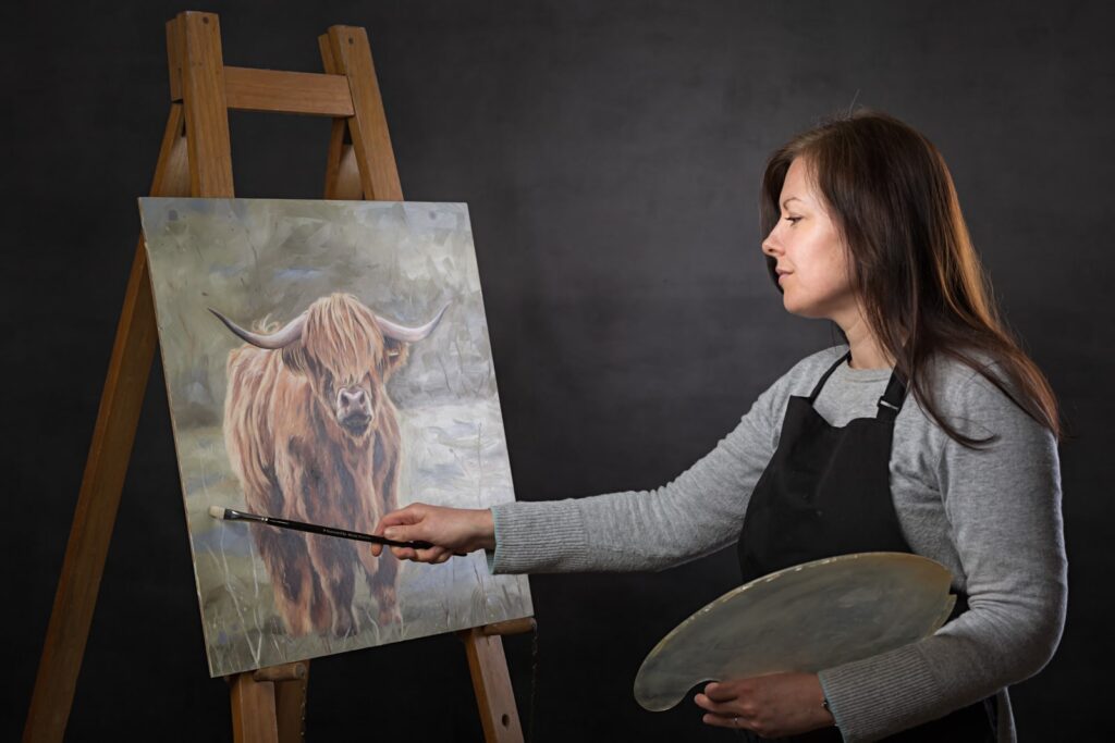 Artist painting a highland cattle portrait on an easel against a dark background, holding a brush and palette.