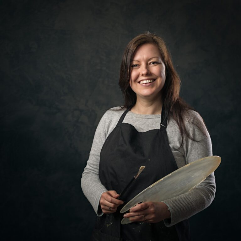 A woman wearing a black apron over a grey cardigan holds a wooden palette and brush, smiling at the camera against a dark background.