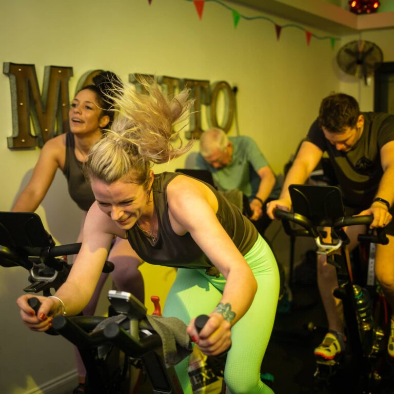 A group of people on stationary bikes in a bright yellow fitness studio during an indoor cycling class.