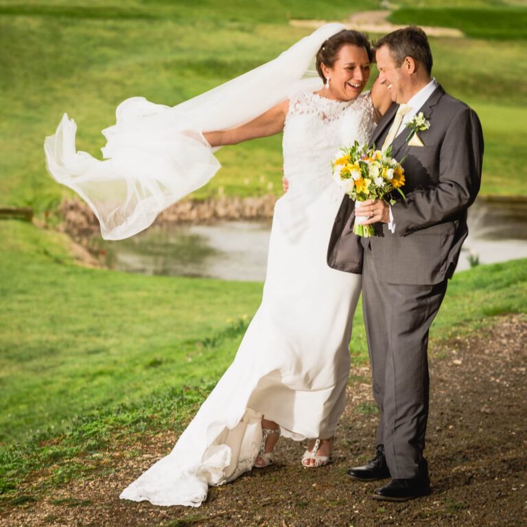 A bride and groom stand together on a country path, smiling at each other. The bride wears a white lace dress with a flowing veil, holding a yellow and white bouquet. The groom wears a grey suit with a buttonhole. Green fields and a stream are visible behind them.
