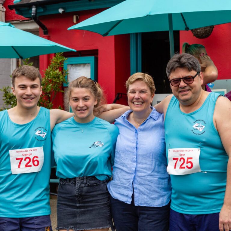 Four people in matching turquoise shirts at a 10K race event, smiling at the camera in front of a red and turquoise building.