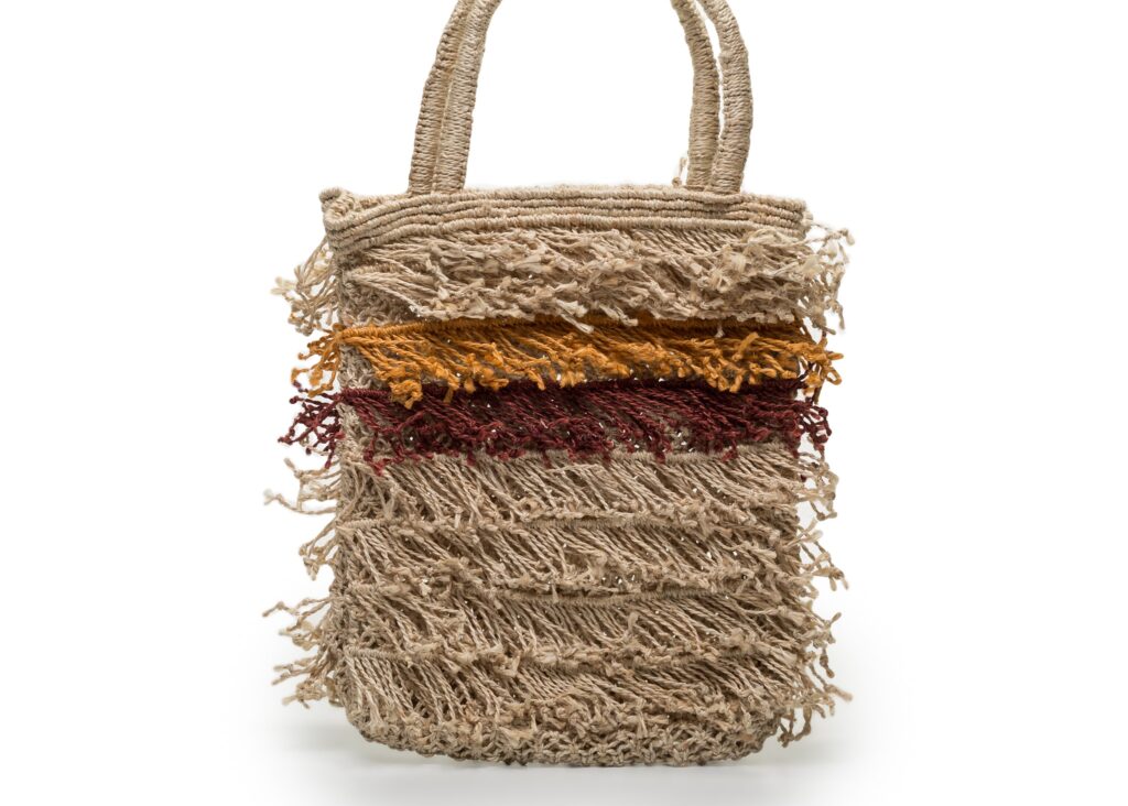 A woven straw handbag with horizontal stripes in cream, gold, burgundy and natural tan, featuring fringed edges and rope handles.