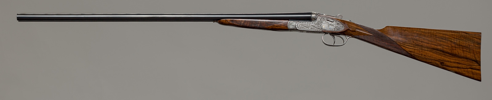 A side-by-side double-barrel shotgun with wooden furniture and ornate engraved silver receiver, displayed against a grey background.