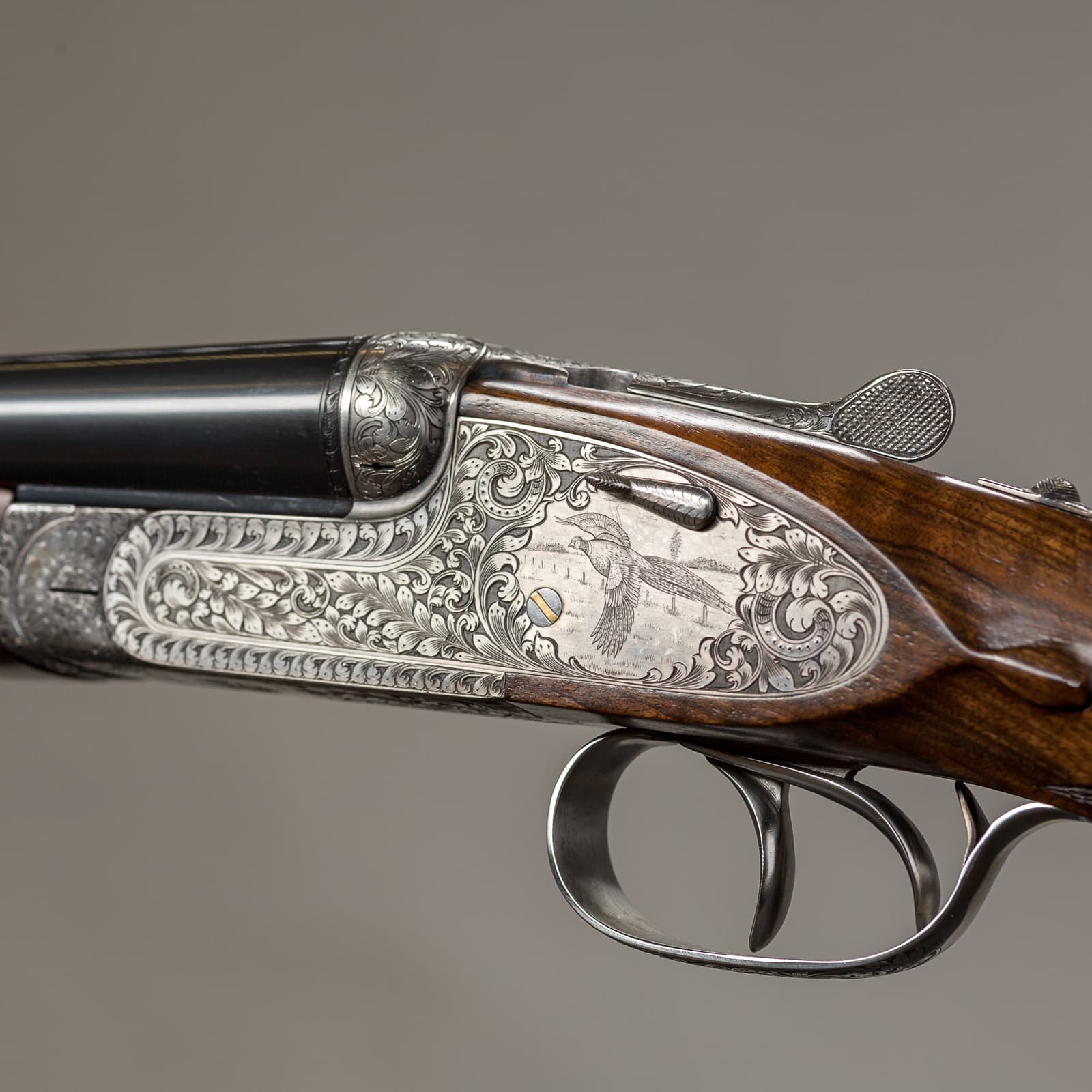 A finely engraved double-barrelled shotgun with ornate scrollwork on the receiver and wood furniture, shown in profile against a neutral grey background.