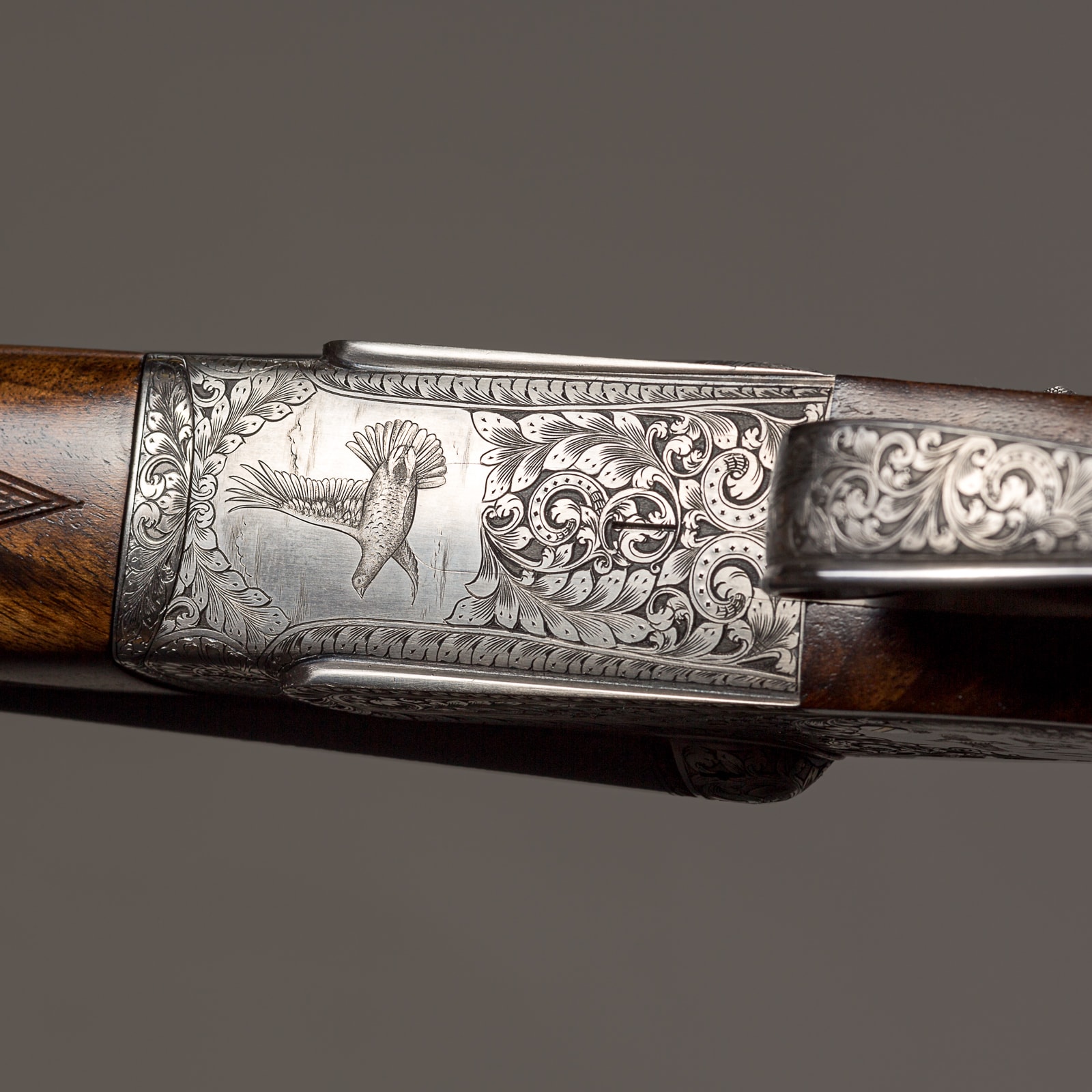 Ornate shotgun receiver with detailed engraved silver metalwork and wood furniture, displayed against a grey background.