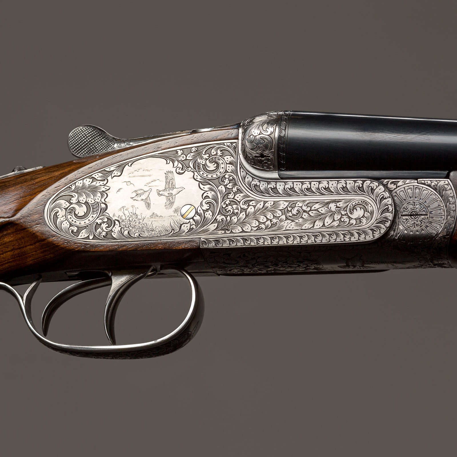 Ornate side-by-side shotgun with intricate silver engraving and wood furniture, shown in profile against a grey background.