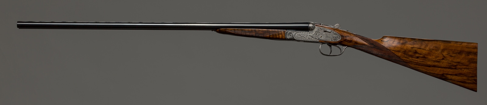 Side view of an ornate over-and-under shotgun with wooden stock against a grey background.