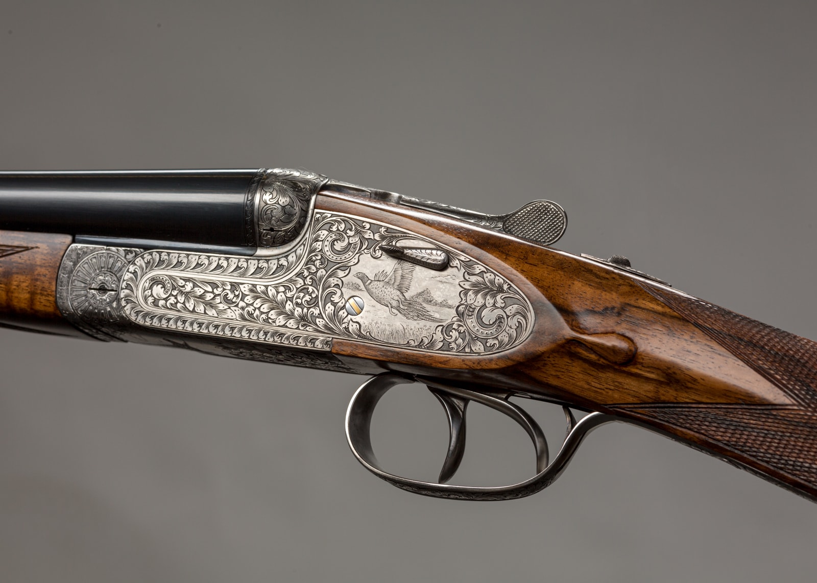 A finely engraved side-by-side shotgun with ornate metalwork and polished wood furniture, displayed against a neutral grey background.