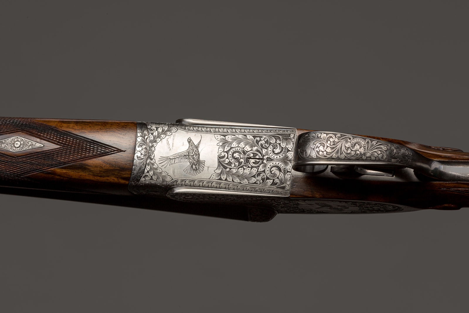 A ornately engraved shotgun receiver with wooden stock, displayed against a grey background.