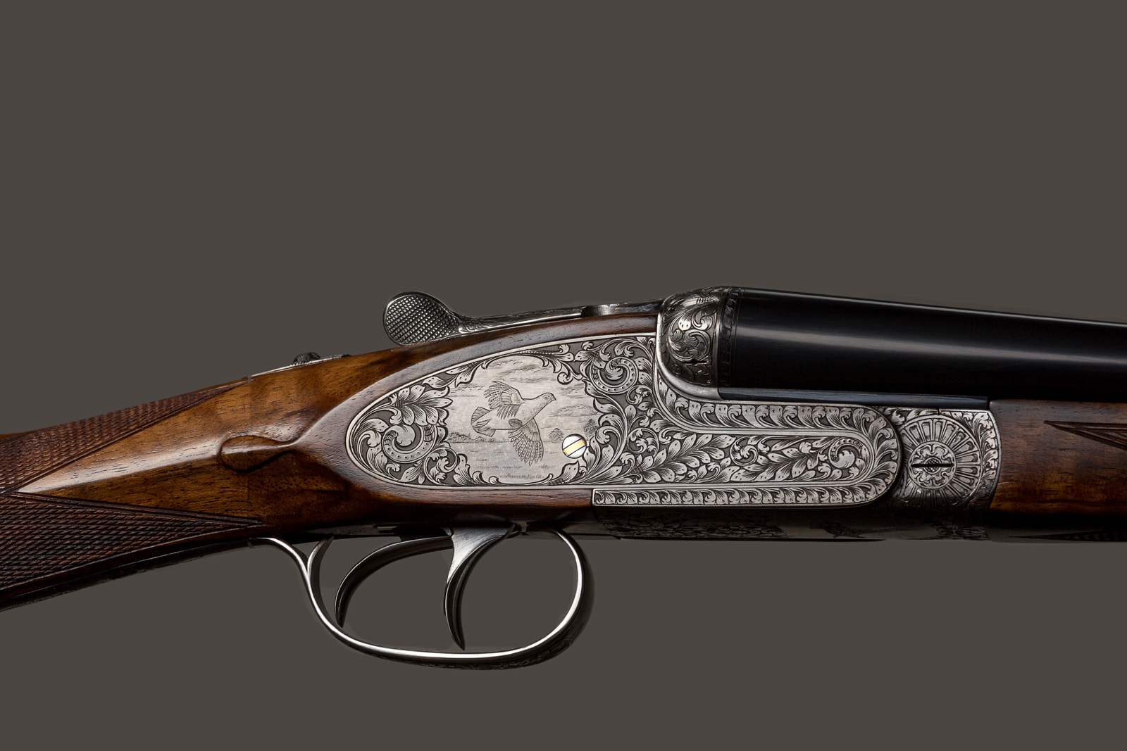 A side-by-side double-barrel shotgun with ornate engraved receiver and polished wood stock, displayed against a neutral grey background.