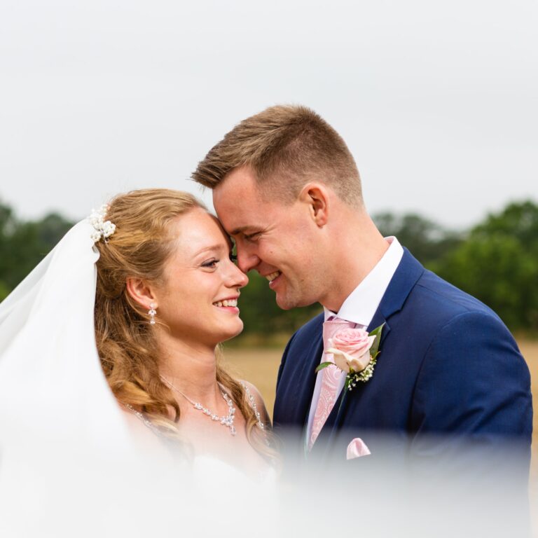 A bride and groom face each other smiling outdoors. She wears a white veil and necklace; he wears a navy suit with a pink rose boutonniere. Trees are visible behind them.