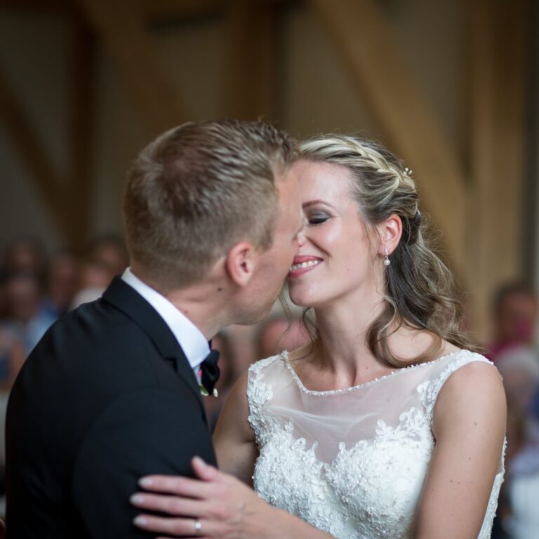 A couple shares a first dance at their wedding. The bride wears an ornate white beaded gown and smiles with eyes closed. Guests watch in the blurred background.