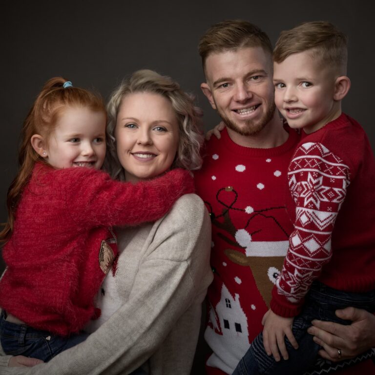 A family of four wearing red festive jumpers smiles at the camera against a dark grey background.