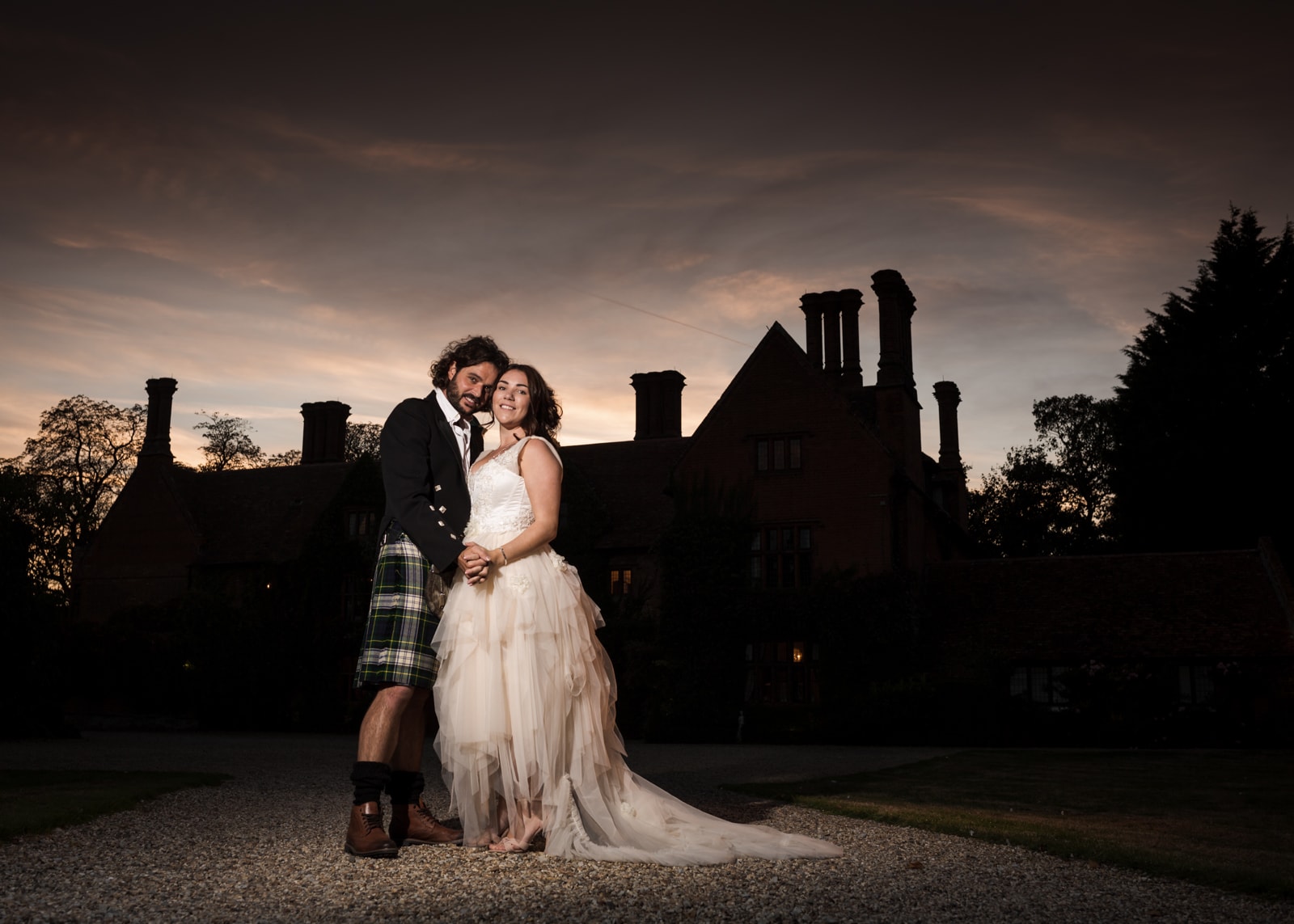 A couple in wedding attire stand together at dusk before a grand manor house silhouetted against the sky.