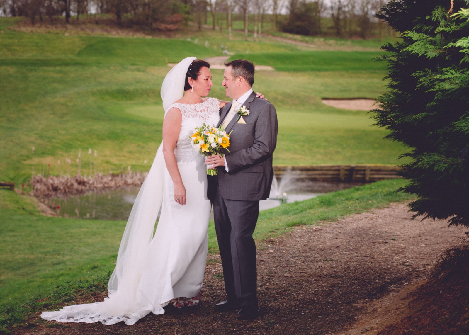 A bride and groom stand together on a garden path, looking at each other. The bride wears a white lace dress and veil, holding a yellow and white bouquet. The groom wears a grey suit. Behind them is a manicured garden with green lawn and a small pond.