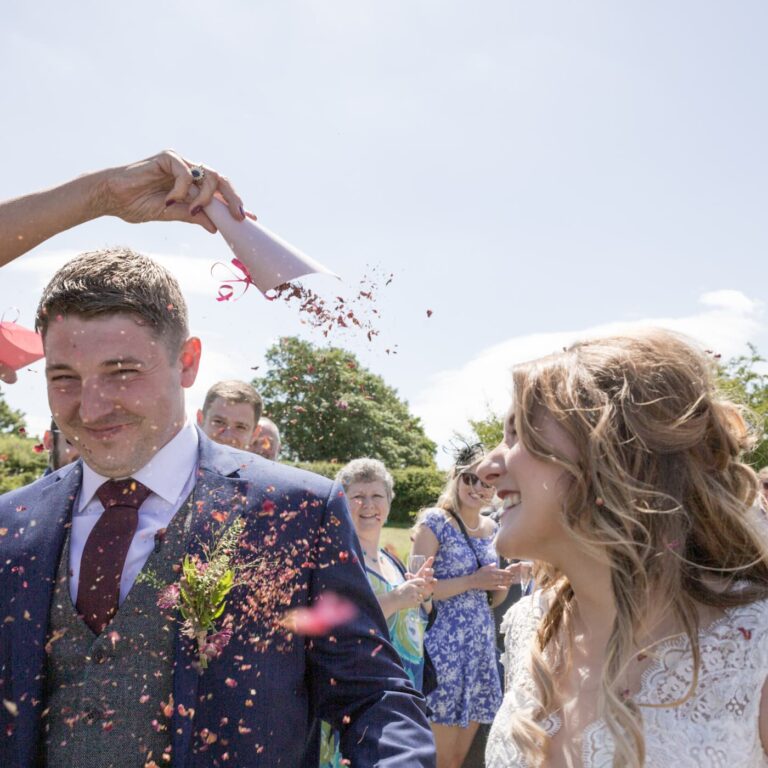 A bride and groom being showered with confetti outdoors on their wedding day, both smiling at each other.