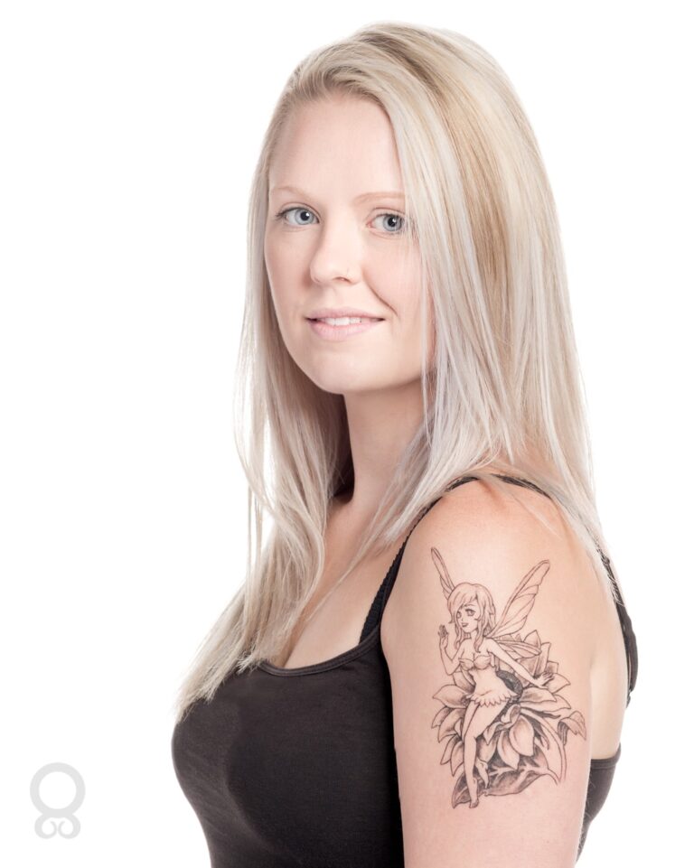 A woman with long pale blonde hair wearing a black sleeveless top, looking over her shoulder. She has a fairy tattoo on her upper arm.