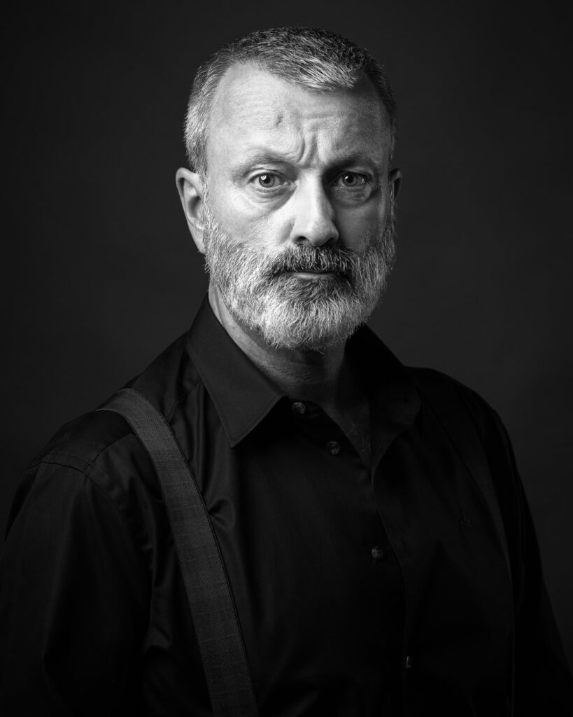 Black and white portrait of a bearded man with grey hair wearing a dark shirt against a dark background, looking directly at camera.